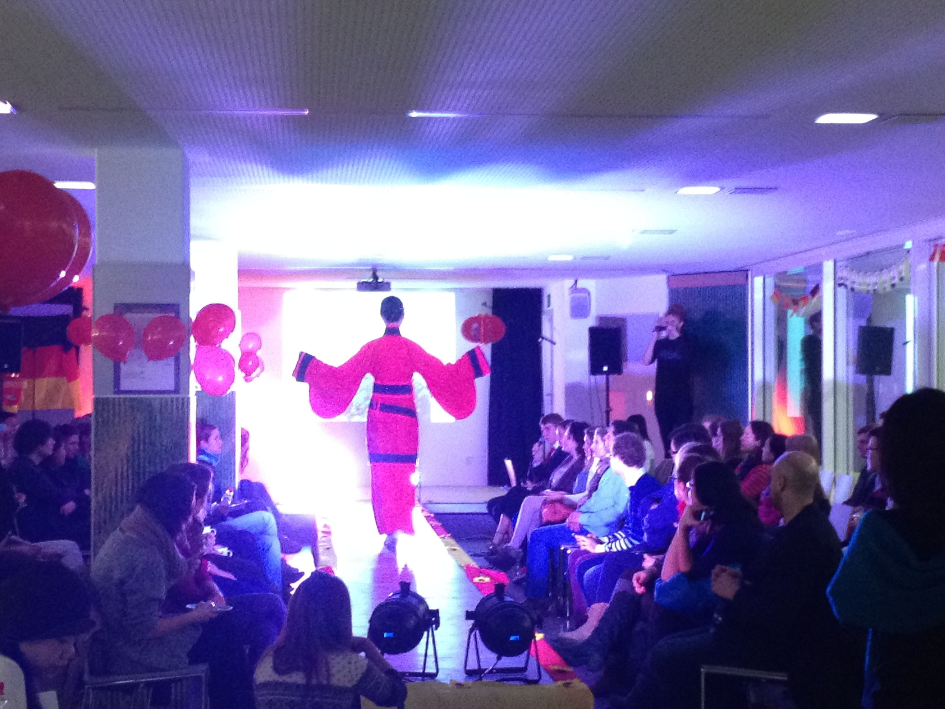 Eine chinesische Modeschau: in der Mitte befindet sich der Laufsteg, auf dem ein Model in einem roten Kimono steht. Rechts und links sitzen die Zuschauer.